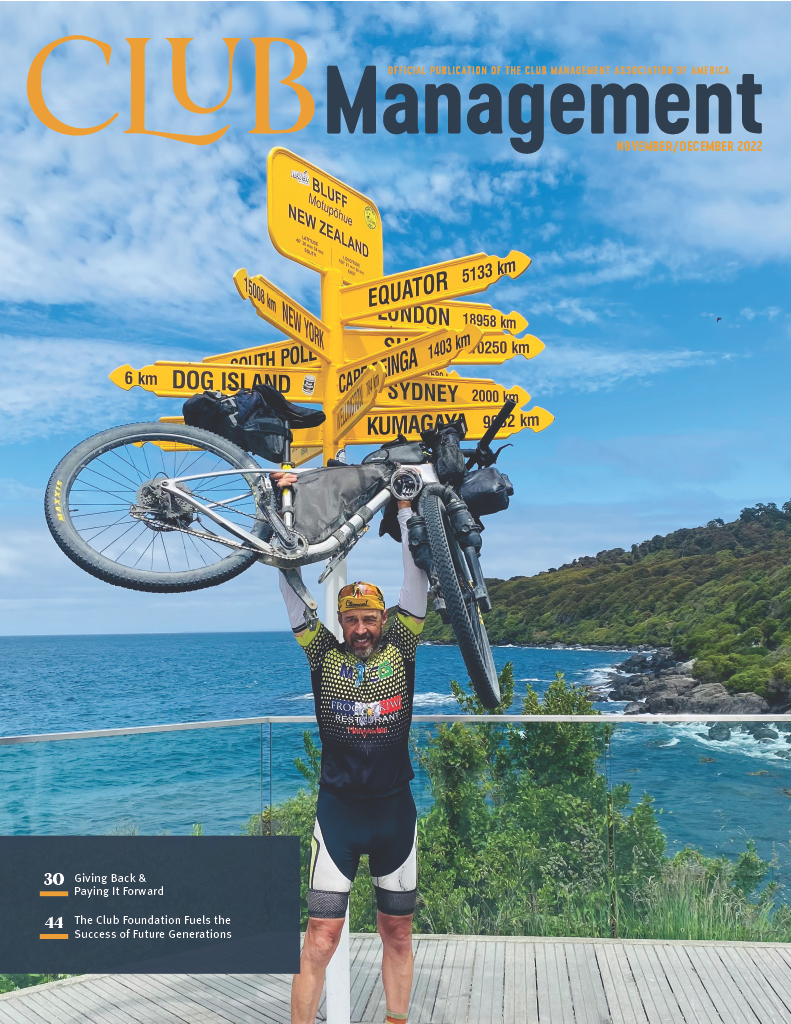 November/December Issue Cover featuring a cyclist holding his bike up in front of a sign pointing out the directions of different locations around the world